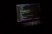 A laptop with an illuminated keyboard displays lines of code on its screen against a dark background. The code is highlighted with various colors to indicate syntax.