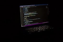 A laptop with an illuminated keyboard displays lines of code on its screen against a dark background. The code is highlighted with various colors to indicate syntax.