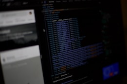 A computer screen displaying a dark-themed text editor with code in various colors. The left side shows the interface of a web inspector or development tool, with some unreadable elements beside the code. The background appears blurred with faint details of another screen or window.