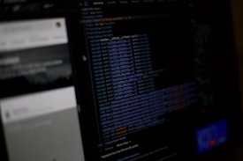 A computer screen displaying a dark-themed text editor with code in various colors. The left side shows the interface of a web inspector or development tool, with some unreadable elements beside the code. The background appears blurred with faint details of another screen or window.