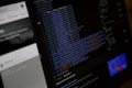 A close-up view of a computer screen displaying HTML or web development code. The text is colored with syntax highlighting, including blue and orange. On the left side, there is a blurry section showing what seems to be a user interface with partially visible text. The overall scene suggests a focus on coding or software development work.