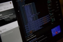 A close-up view of a computer screen displaying HTML or web development code. The text is colored with syntax highlighting, including blue and orange. On the left side, there is a blurry section showing what seems to be a user interface with partially visible text. The overall scene suggests a focus on coding or software development work.