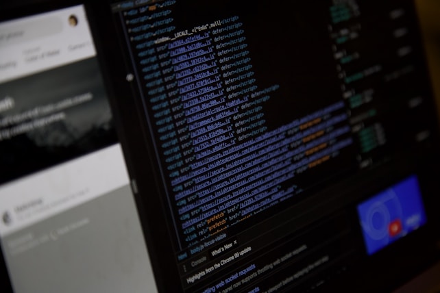 A close-up view of a computer screen displaying HTML or web development code. The text is colored with syntax highlighting, including blue and orange. On the left side, there is a blurry section showing what seems to be a user interface with partially visible text. The overall scene suggests a focus on coding or software development work.