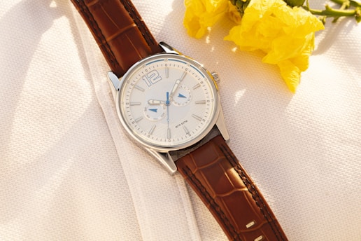 A wristwatch with a silver dial and brown leather strap is placed on a textured white surface next to vibrant yellow flowers. The watch has blue highlights on the face with two subdials.