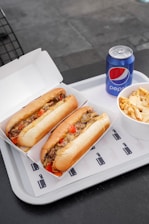 Two sandwiches filled with meat, cheese, and vegetables are placed in open cardboard boxes on a white tray. Next to them, there is a bowl containing fries topped with cheese sauce and a can of Pepsi.