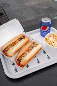 Two sandwiches filled with meat, cheese, and vegetables are placed in open cardboard boxes on a white tray. Next to them, there is a bowl containing fries topped with cheese sauce and a can of Pepsi.