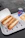 Two sandwiches filled with meat, cheese, and vegetables are placed in open cardboard boxes on a white tray. Next to them, there is a bowl containing fries topped with cheese sauce and a can of Pepsi.