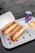 Two sandwiches filled with meat, cheese, and vegetables are placed in open cardboard boxes on a white tray. Next to them, there is a bowl containing fries topped with cheese sauce and a can of Pepsi.