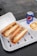 Two sandwiches filled with meat, cheese, and vegetables are placed in open cardboard boxes on a white tray. Next to them, there is a bowl containing fries topped with cheese sauce and a can of Pepsi.