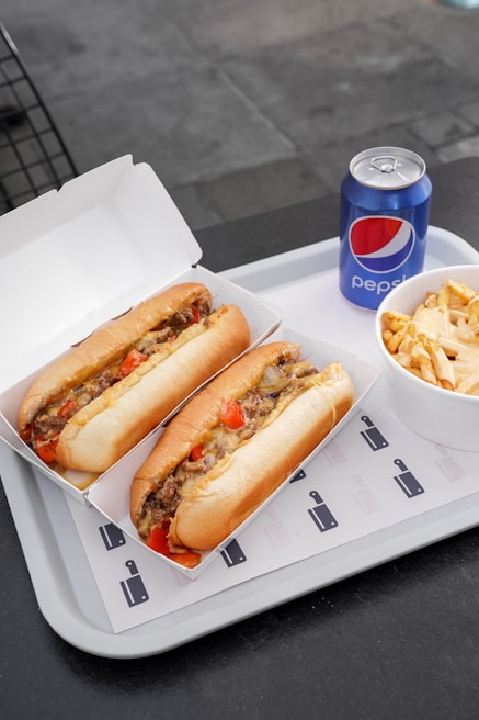 Two sandwiches filled with meat, cheese, and vegetables are placed in open cardboard boxes on a white tray. Next to them, there is a bowl containing fries topped with cheese sauce and a can of Pepsi.