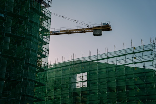 A construction site with a prominent crane extending over the partially covered building structure. The building is wrapped in green safety mesh, and scaffolding surrounds it, indicating ongoing construction work. The sky is clear, suggesting daytime.