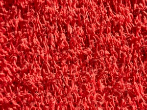 The image features a close-up view of a red textured surface, possibly a mat or carpet, with a dense pattern of loops and fibers.