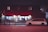 A building with a store called 'Youniform Market' features prominently, displaying a red awning with text. Brightly lit window displays show clothes and mannequins inside. A white minivan is parked in front of the store on a quiet street at night, with some streetlights providing illumination.
