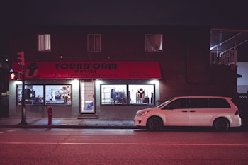 A building with a store called 'Youniform Market' features prominently, displaying a red awning with text. Brightly lit window displays show clothes and mannequins inside. A white minivan is parked in front of the store on a quiet street at night, with some streetlights providing illumination.