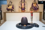 A spiritual setting featuring three ornate statues dressed in vibrant purple and gold garments. In front of them is a large, smooth black sculpture with a small coconut at its base and a red powder mark on top. A trident adorned with a red cloth and prayer beads stands beside the sculpture.