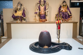 A spiritual setting featuring three ornate statues dressed in vibrant purple and gold garments. In front of them is a large, smooth black sculpture with a small coconut at its base and a red powder mark on top. A trident adorned with a red cloth and prayer beads stands beside the sculpture.