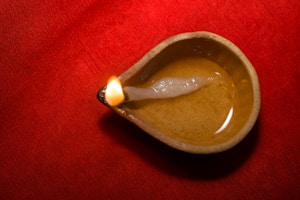 A traditional oil lamp, made of clay, rests on a vibrant red fabric surface. The lamp contains a small amount of oil and a wick, which is lit, producing a soft, warm glow.