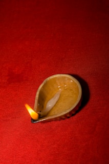 A small ceramic oil lamp with a flame burning at the wick, placed against a rich red fabric background. The lamp, shaped like a traditional diya, is made of a glossy material with visible textures and soft light reflecting off its surface.