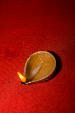 A small ceramic oil lamp with a flame burning at the wick, placed against a rich red fabric background. The lamp, shaped like a traditional diya, is made of a glossy material with visible textures and soft light reflecting off its surface.