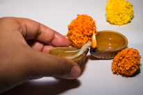 Hands gently pouring Shree Aradhana pooja oil into a temple lamp surrounded by marigold flowers.