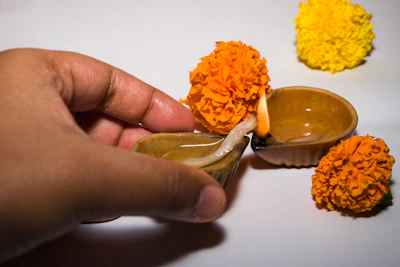 Hands gently pouring Shree Aradhana pooja oil into a temple lamp surrounded by marigold flowers.