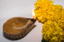 A traditional clay oil lamp with a wick is lit, casting a soft flame. Bright yellow marigold flowers are placed beside it on a plain background, suggesting a festive or ritualistic setting.