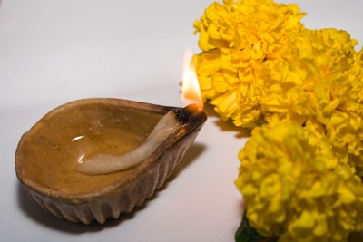 A traditional clay oil lamp with a wick is lit, casting a soft flame. Bright yellow marigold flowers are placed beside it on a plain background, suggesting a festive or ritualistic setting.