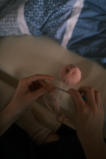 Hands are engaging in crocheting with pink yarn, accompanied by a blue crocheting needle. A ball of pink yarn lies on a surface nearby, and there is blue-patterned fabric in the background.
