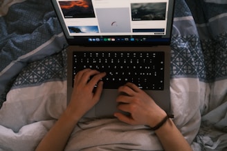 A pair of hands is using a laptop on a patterned blanket, browsing images on a website. The lighting suggests a cozy, indoor atmosphere.