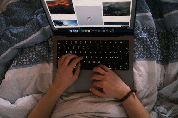 A pair of hands is using a laptop on a patterned blanket, browsing images on a website. The lighting suggests a cozy, indoor atmosphere.