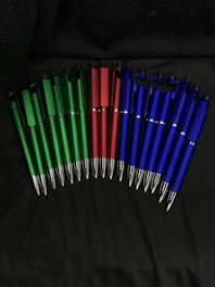 A collection of pens arranged in a semi-circle on a dark surface, with the pens grouped by color: green, red, and blue.
