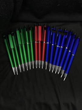 A collection of pens arranged in a semi-circle on a dark surface, with the pens grouped by color: green, red, and blue.