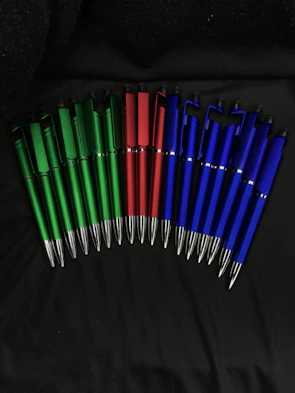 A collection of ballpoint pens in various colors laid out on a wooden desk.