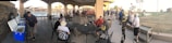 A group of people gathered under a large covered patio area near a pool. Some individuals are seated around tables, while others stand near open coolers. The setting appears social, with many wearing casual sportswear.