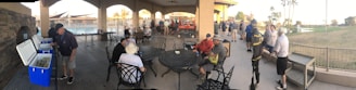 A group of people gathered under a large covered patio area near a pool. Some individuals are seated around tables, while others stand near open coolers. The setting appears social, with many wearing casual sportswear.