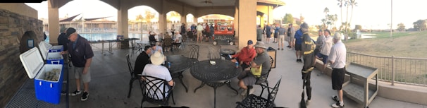 A group of people gathered under a large covered patio area near a pool. Some individuals are seated around tables, while others stand near open coolers. The setting appears social, with many wearing casual sportswear.
