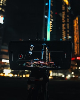 A smartphone is mounted on a tripod, capturing a skyline with illuminated skyscrapers. The screen displays a vivid image of the cityscape at night, with various bright lights and buildings. Surrounding the phone, the background blurs into warm glowing bokeh effects, enhancing the nighttime ambiance.