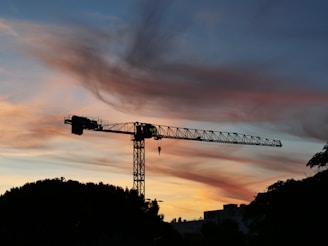A towering crane silhouetted against a vibrant sunset over a busy urban construction site.