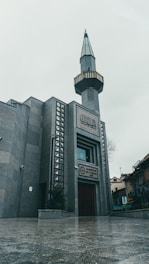 A modern mosque with a tall minaret and geometric architectural designs. The building features grey stone walls and intricate patterns around the entrance. The sky is overcast, giving a moody atmosphere to the scene.