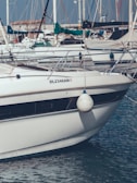 A white boat is docked at a marina, with many other boats visible in the background. The boat has a fender hanging on the side and identifiable registration DL2346AN. There are numerous masts and sails in the surrounding area.