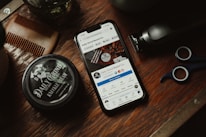 A wooden surface is adorned with grooming tools including a comb, a pair of scissors, a trimmer, and a jar labeled 'Daily Grind Knuckle Cream.' A smartphone displaying a social media page for 'Daily Grind Beard Co.' is centrally placed.