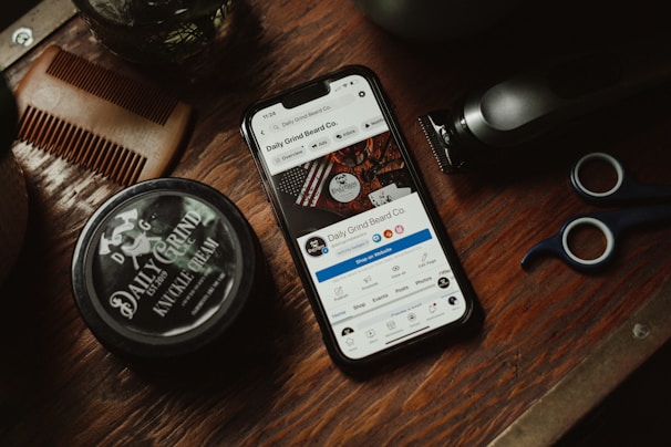 A wooden surface is adorned with grooming tools including a comb, a pair of scissors, a trimmer, and a jar labeled 'Daily Grind Knuckle Cream.' A smartphone displaying a social media page for 'Daily Grind Beard Co.' is centrally placed.