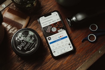 A wooden surface is adorned with grooming tools including a comb, a pair of scissors, a trimmer, and a jar labeled 'Daily Grind Knuckle Cream.' A smartphone displaying a social media page for 'Daily Grind Beard Co.' is centrally placed.