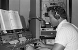 A man wearing headphones operates a vintage radio control panel. He is speaking into a classic microphone while adjusting the dials. Paper scripts are propped up in front of him, and an old reel-to-reel tape machine is visible in the background.