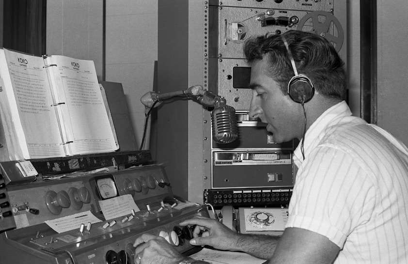 A man wearing headphones operates a vintage radio control panel. He is speaking into a classic microphone while adjusting the dials. Paper scripts are propped up in front of him, and an old reel-to-reel tape machine is visible in the background.