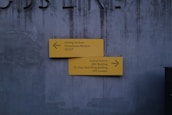 A gray concrete wall serves as the backdrop for two yellow directional signs. The top sign points left, indicating directions to Darling Harbour, Powerhouse Museum, and SICEEP. The bottom sign points right, leading to Central Station, ABC Building, Dr Chau Chak Wing Building, and UTS Campus. The wall has a slightly worn texture and faint letters partially visible at the top.