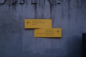 A gray concrete wall serves as the backdrop for two yellow directional signs. The top sign points left, indicating directions to Darling Harbour, Powerhouse Museum, and SICEEP. The bottom sign points right, leading to Central Station, ABC Building, Dr Chau Chak Wing Building, and UTS Campus. The wall has a slightly worn texture and faint letters partially visible at the top.
