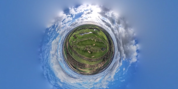 A spherical panorama resembling a tiny planet effect shows a green landscape with fields and pathways surrounded by a vivid blue sky with scattered clouds. The bright light in the sky suggests it is daytime.