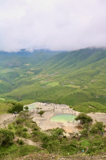 A vast, lush green mountain landscape stretches under a cloudy sky. In the foreground, a natural rock formation featuring small turquoise pools is visible. People are gathered around and in these pools, indicating a popular tourist site.