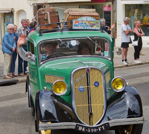 A vintage green car with a classic design is driving down a street. The car has luggage and birdcages on the roof, indicating a journey or road trip. People are gathered on the sidewalk, some waving, suggesting a festive or parade-like atmosphere. The sign on the car's roof reads 'Tour de Bretagne'.
